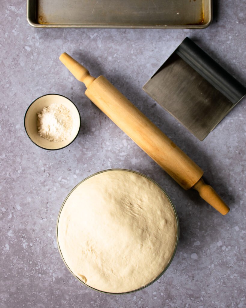 A bowl of dough, a bowl of flour, a rolling pin, a bench scraper and a quarter sheet pan on a counter.