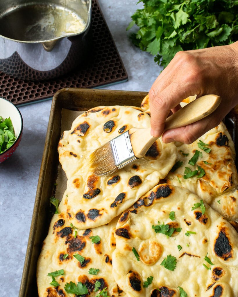 Naan bread on a quarter sheet pan with cilantro sprinkled on top and a hand basting with butter.