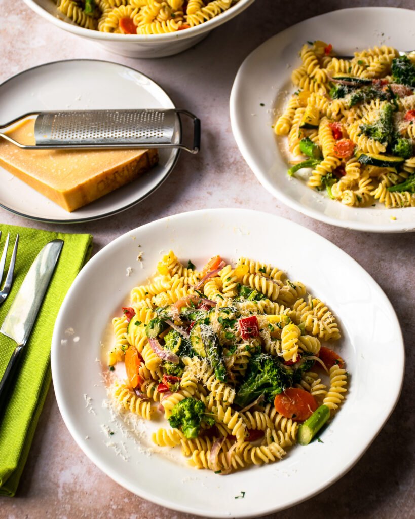 Pasta primavera in a two white bowls with parmesan cheese on a small plate.