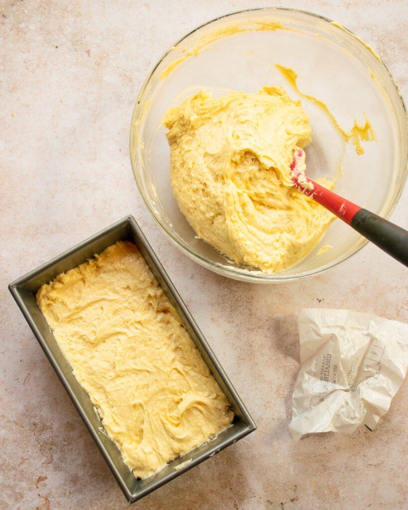 A loaf pan filled with pound cake batter and a bowl with more batter, a spatula and a butter wrapper.