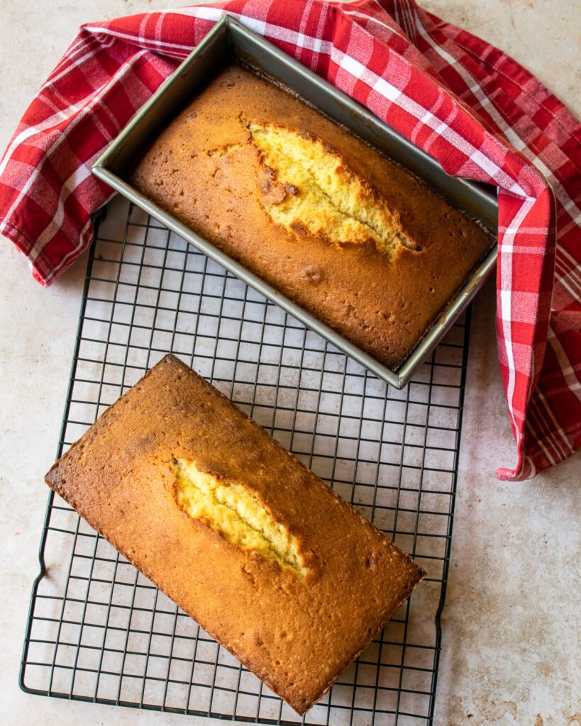 Two pound cakes on a cooling rack - one in a loaf pan and the other out - and a red towel.