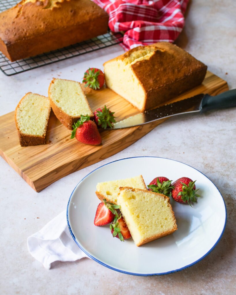 A piece of pound cake on a plate with berries and a whole pound cake loaf in the background.