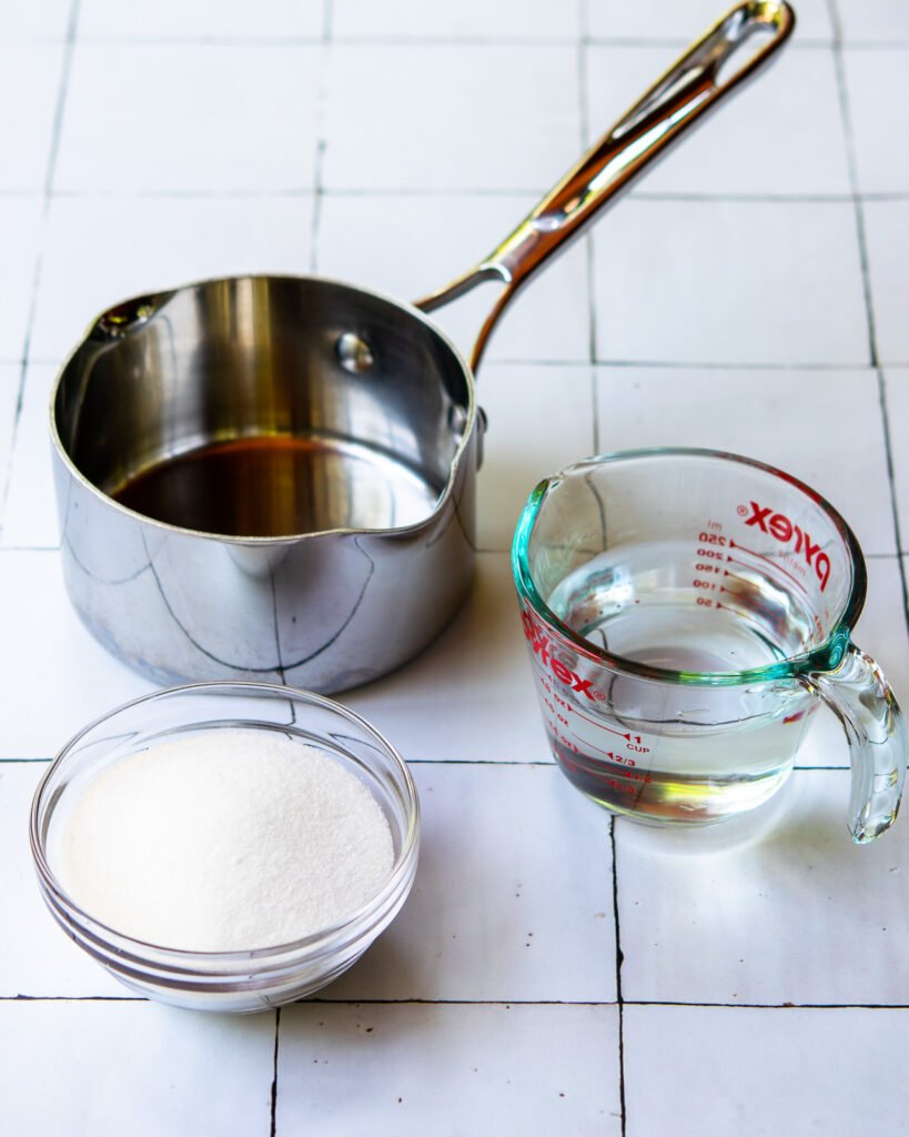 A saucepan, a bowl of sugar and a measuring cup of water on a tiled counter.