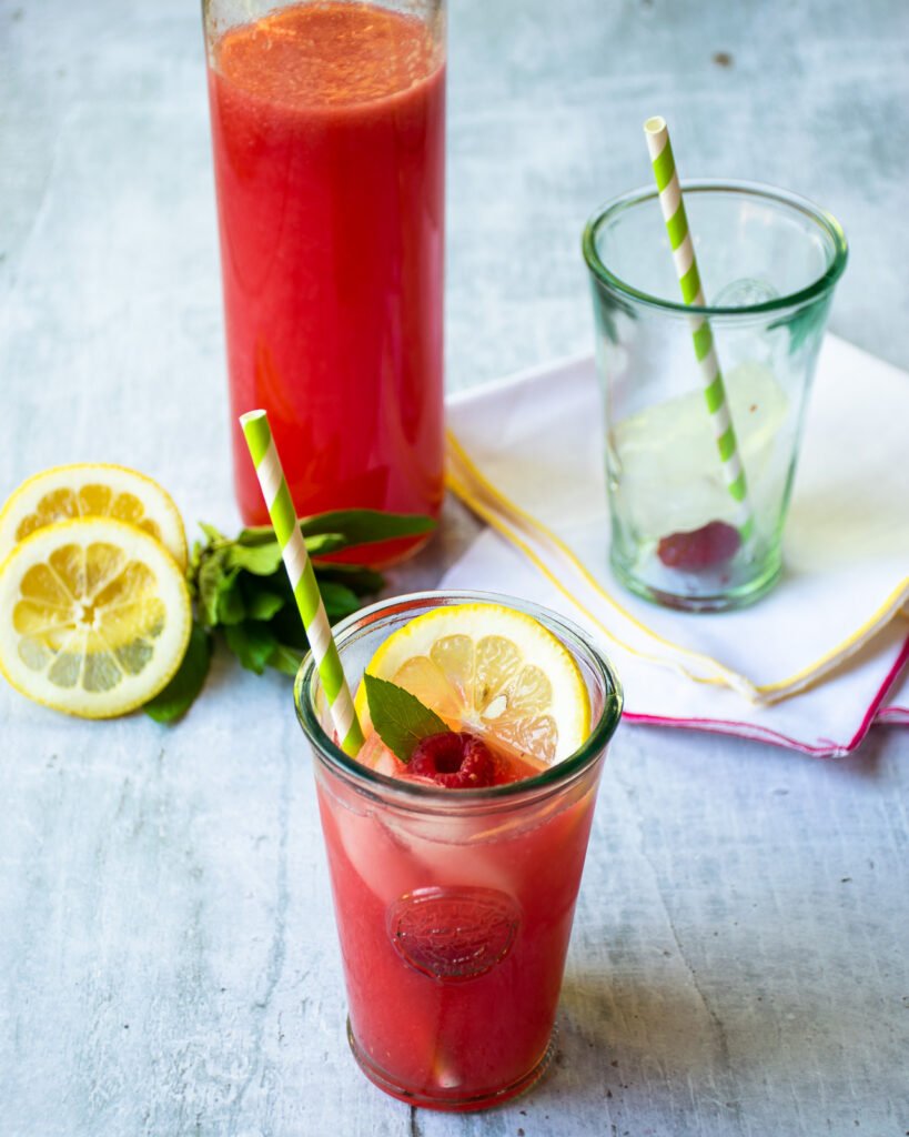 Sparkling raspberry lemonade in a bottle and in a glass with a circle of lemon, a raspberry and a mint leaf.