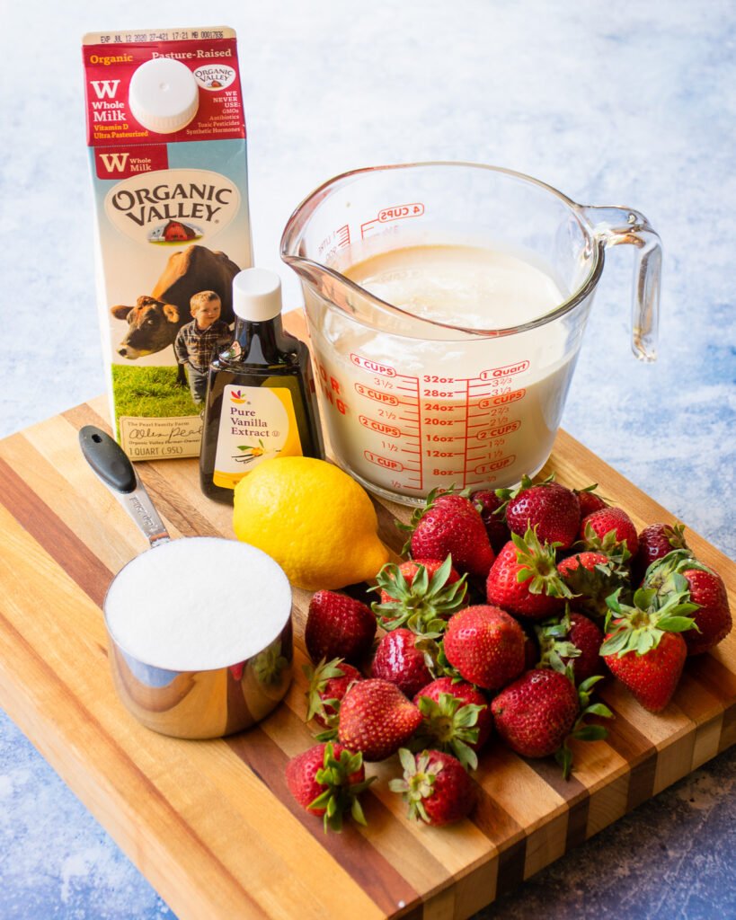 Ingredients on a wooden cutting board - strawberries, sugar, a lemon, cream, milk and vanilla extract.