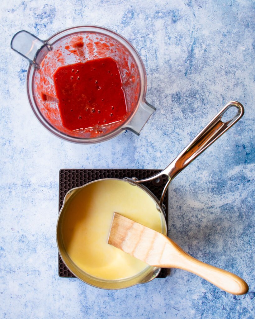 A saucepan with cream and a wooden spoon sitting next to a blender with pureed strawberries.