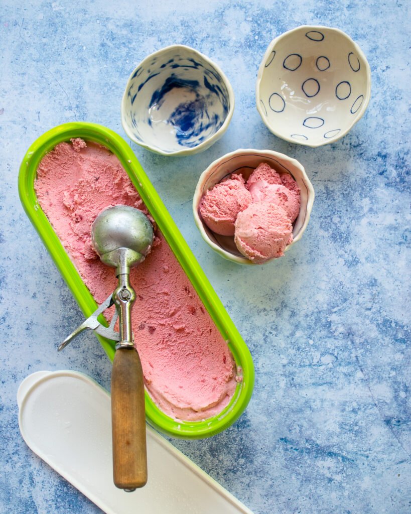 An old fashioned ice cream scoop scooping strawberry ice cream out of a tub into three small bowls on a blue countertop.
