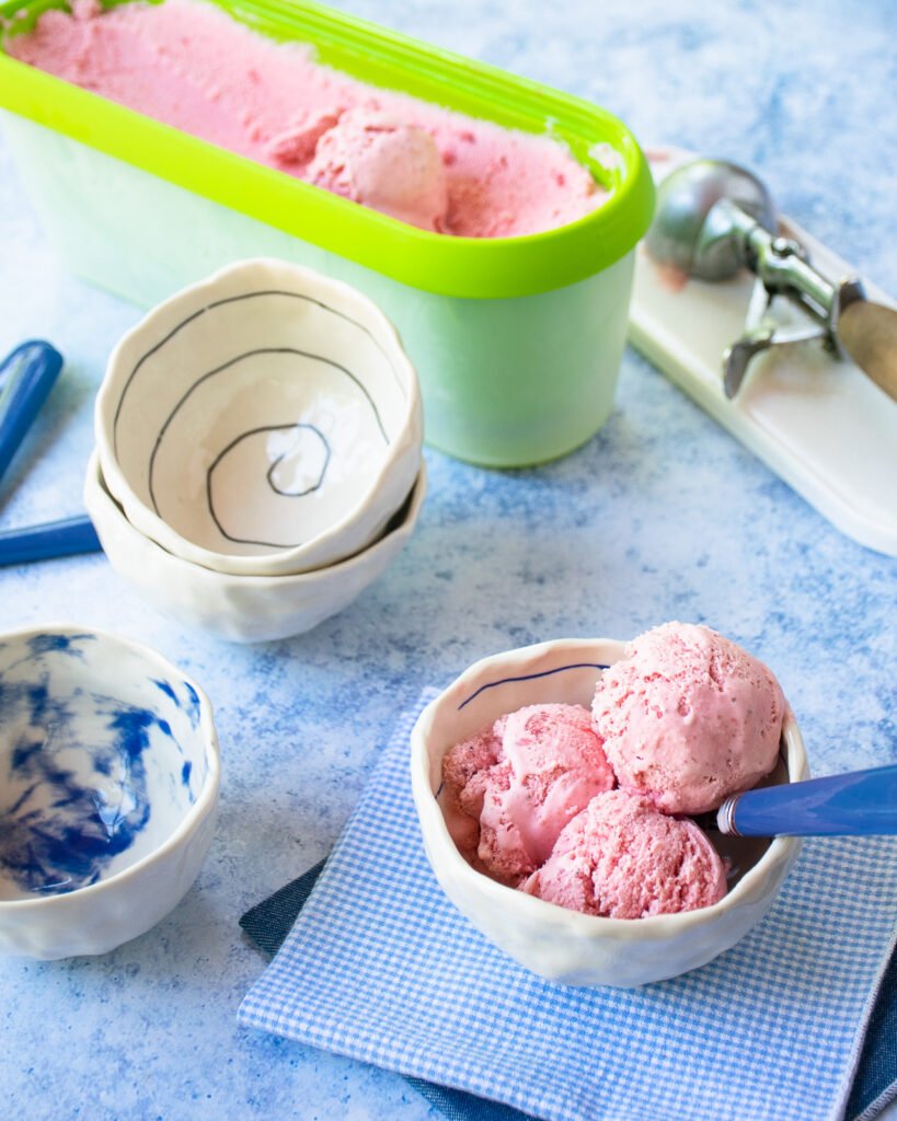 Three scoops of ice cream in a small white bowl with bowls, spoons, a scoop and a tub of ice cream in the background.
