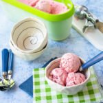 A bowl of ice cream on a green and white checkered napkin with an ice cream tub and more bowls in the background.