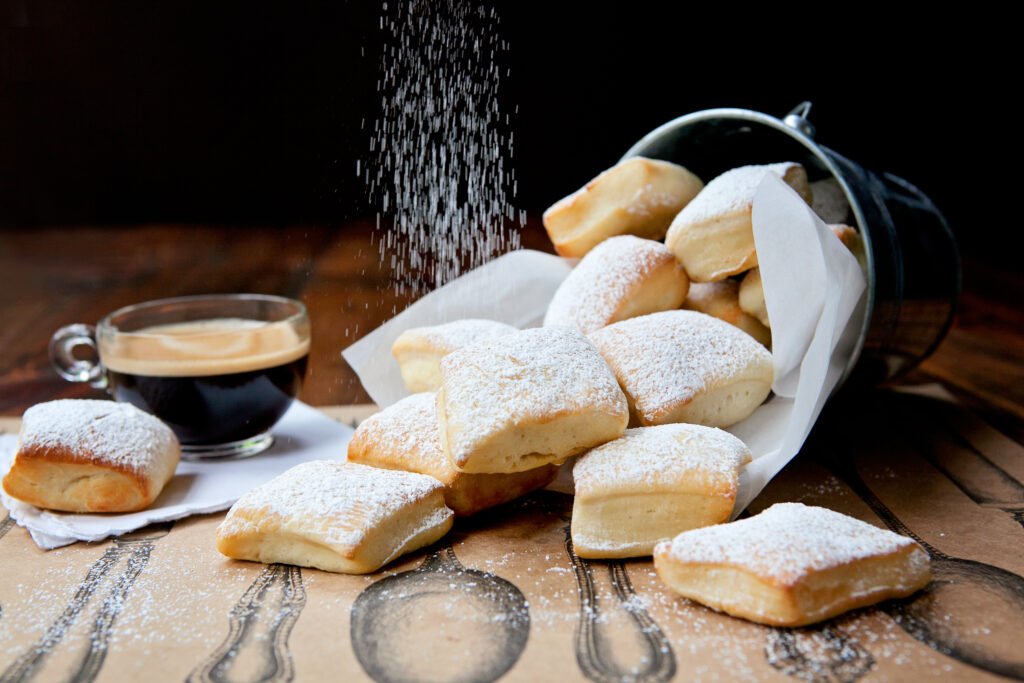 A bucket of beignets tipped over onto a table with a cup of coffee in the background and powdered sugar dusting down from above.