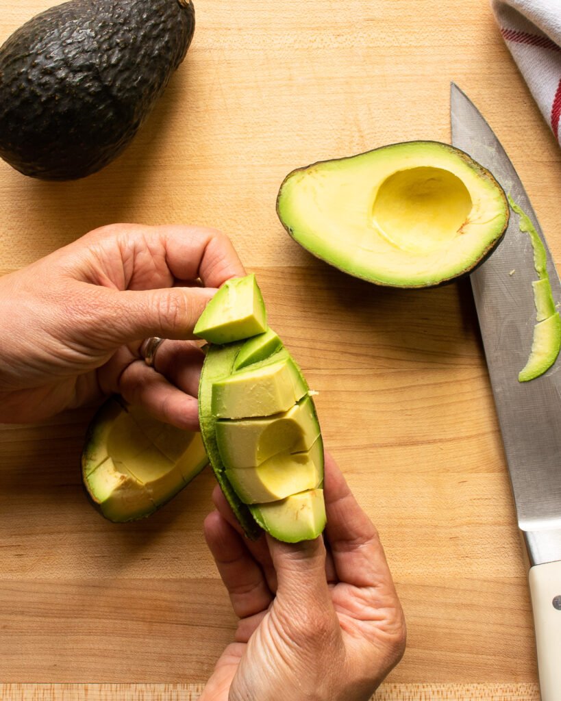 Two hands peeling the peel of avocado away from diced flesh.