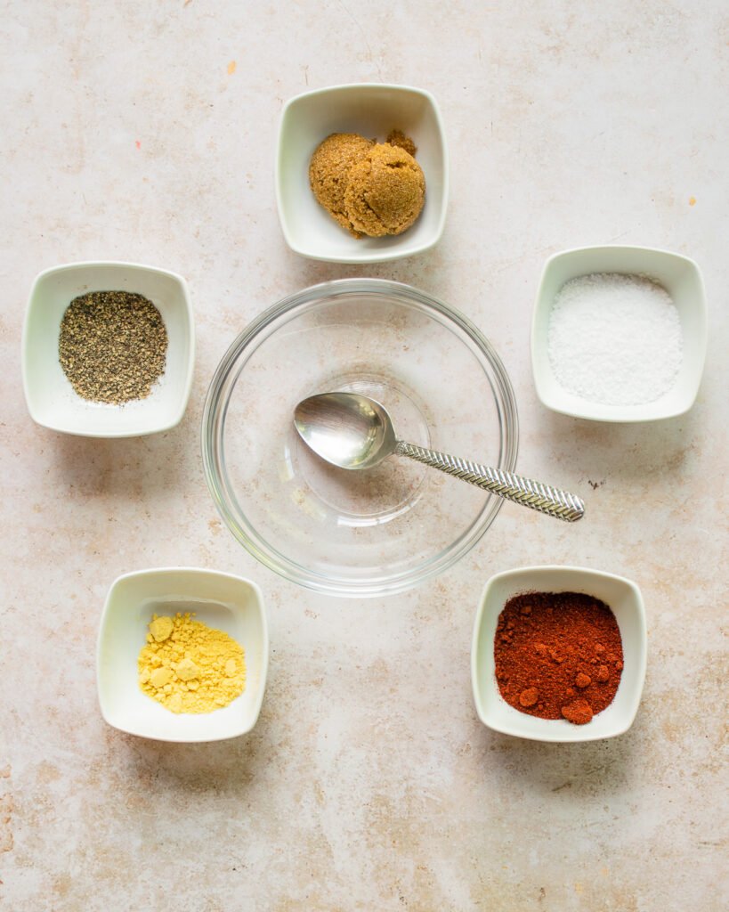 Five white dishes with spices in them and an empty glass bowl with spoon in the center.