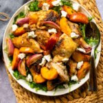 Chicken and Baked Peach Sheet Pan Dinner on a white platter with serving spoons, all on a rattan placemat.