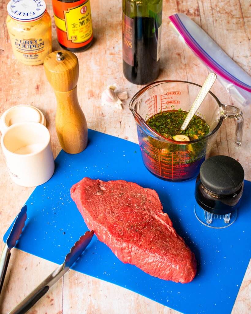 A raw top round steak on a blue cutting board with ingredients and a needle tenderizer near by.