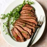 Grilled London Broil, sliced on a white oval platter with fresh thyme and a serving fork.
