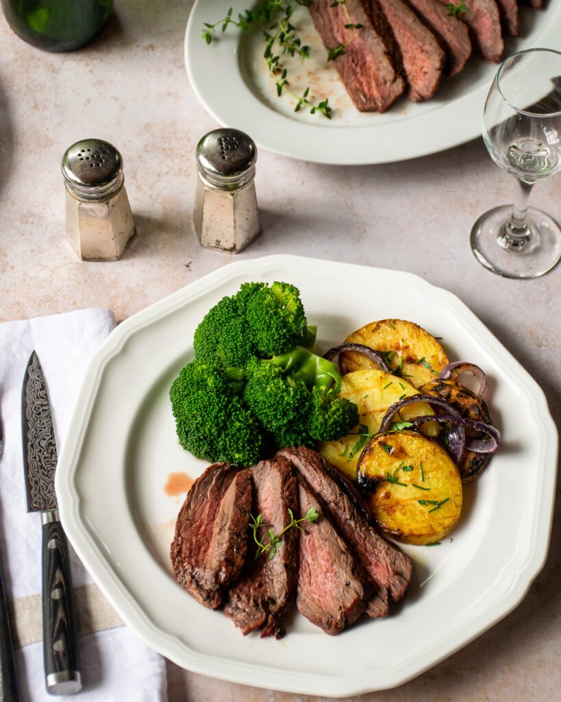 A white dinner plate with grilled marinated London broil, broccoli and grilled potatoes with more steak on a platter in the background.