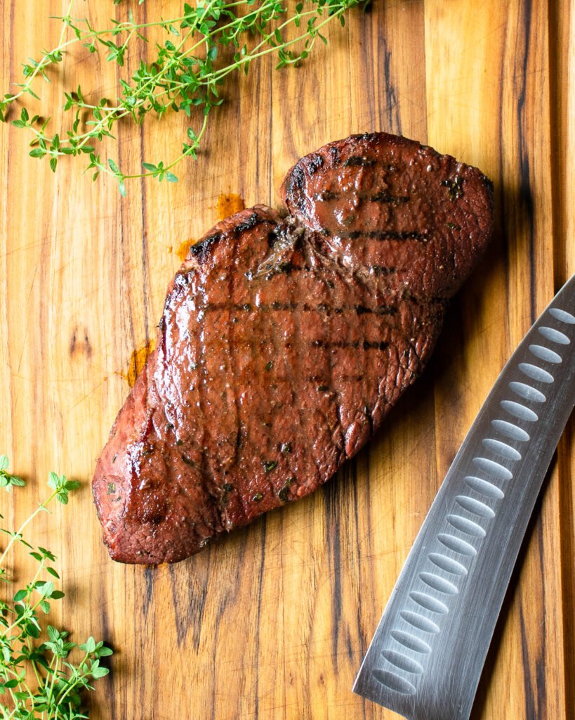 A grilled marinated London broil on a cutting board with a knife.