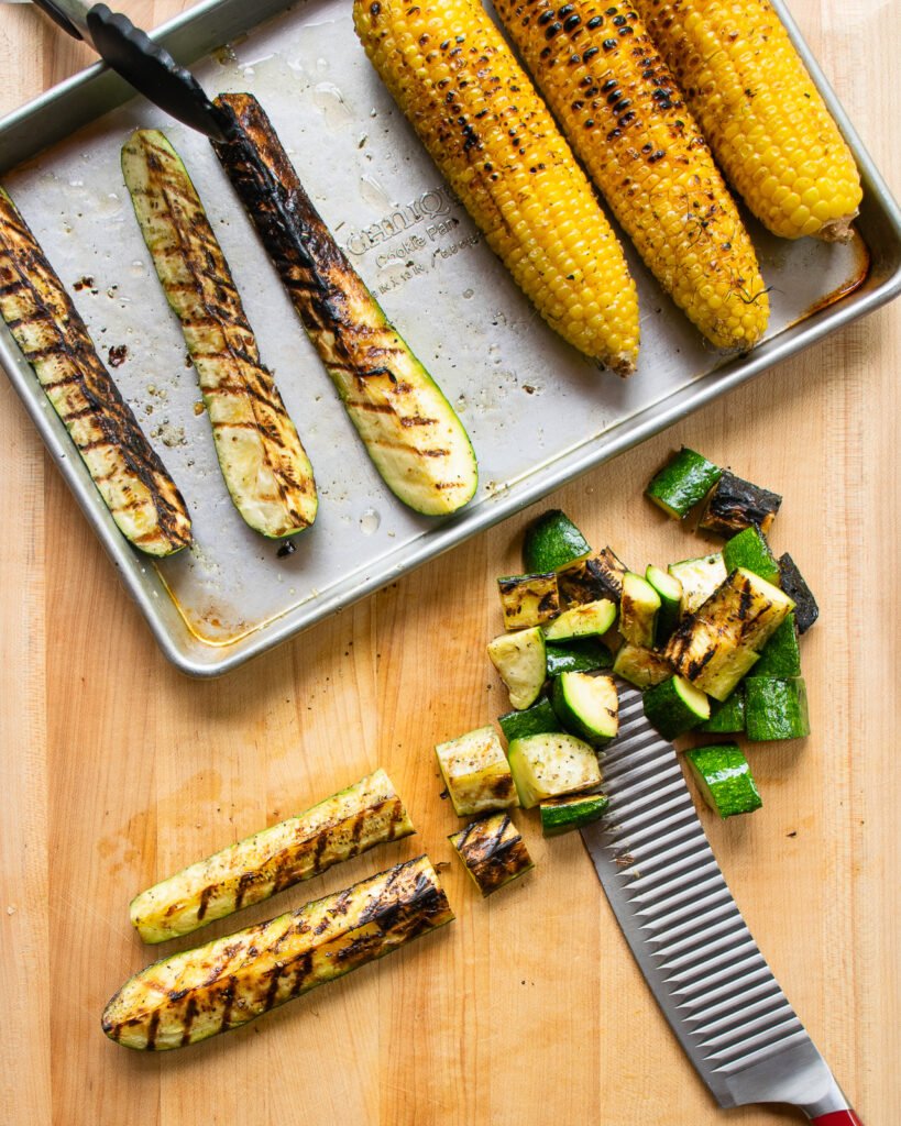 Grilled corn and zucchini on a quarter sheet pan next to some cut up zucchini with a knife.