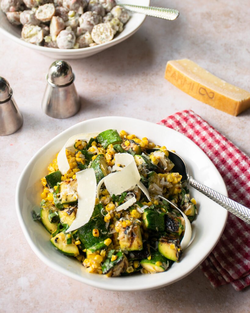 Grilled Parmesan Corn and Zucchini in a white bowl on a table with potato salad in the background.