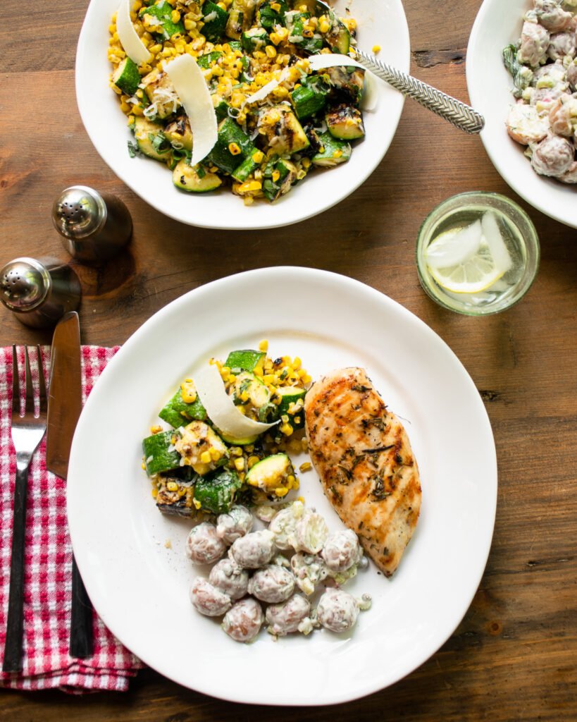 A dinner plate with a grilled chicken breast, some potato salad and some zucchini and corn with a red and white checkered napkin, salt and pepper and a glass of water.
