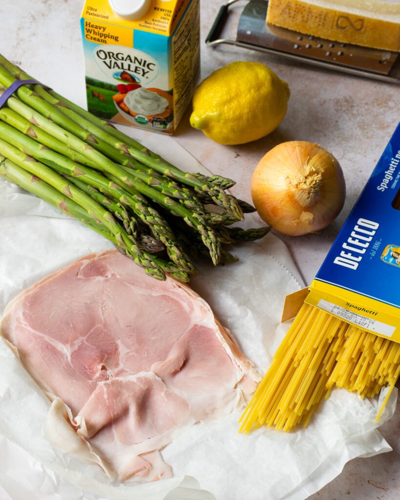 Ingredients on a counter - ham, pasta, asparagus, an onion, lemon, heavy cream and Parmesan cheese.