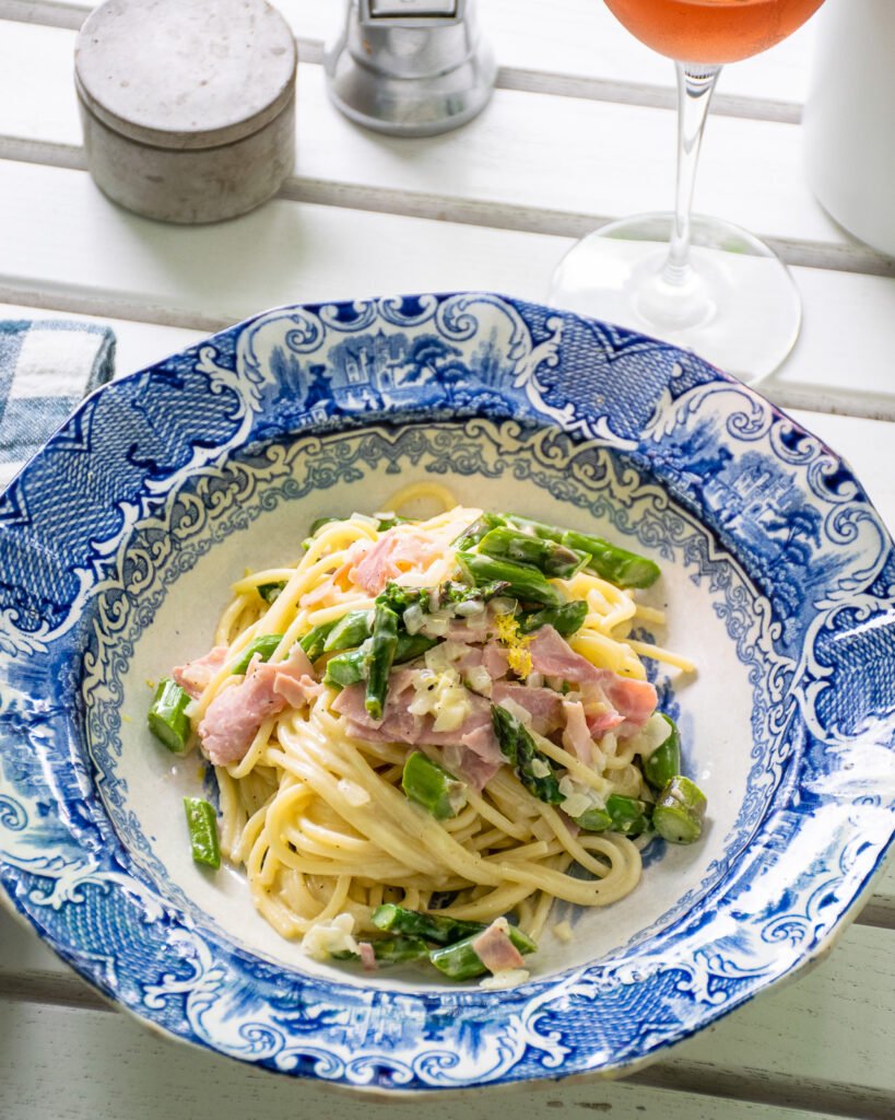 Creamy ham and asparagus pasta in a blue and white bowl on a white wooden table, a glass of rose wine, salt and pepper.