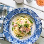 Creamy ham and asparagus pasta in a blue and white bowl on a white wooden table with a glass of rose wine, salt and pepper.