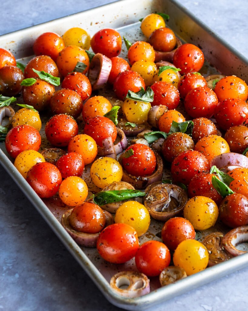 Cherry tomatoes on a sheet pan with seasonings, shallots and basil.