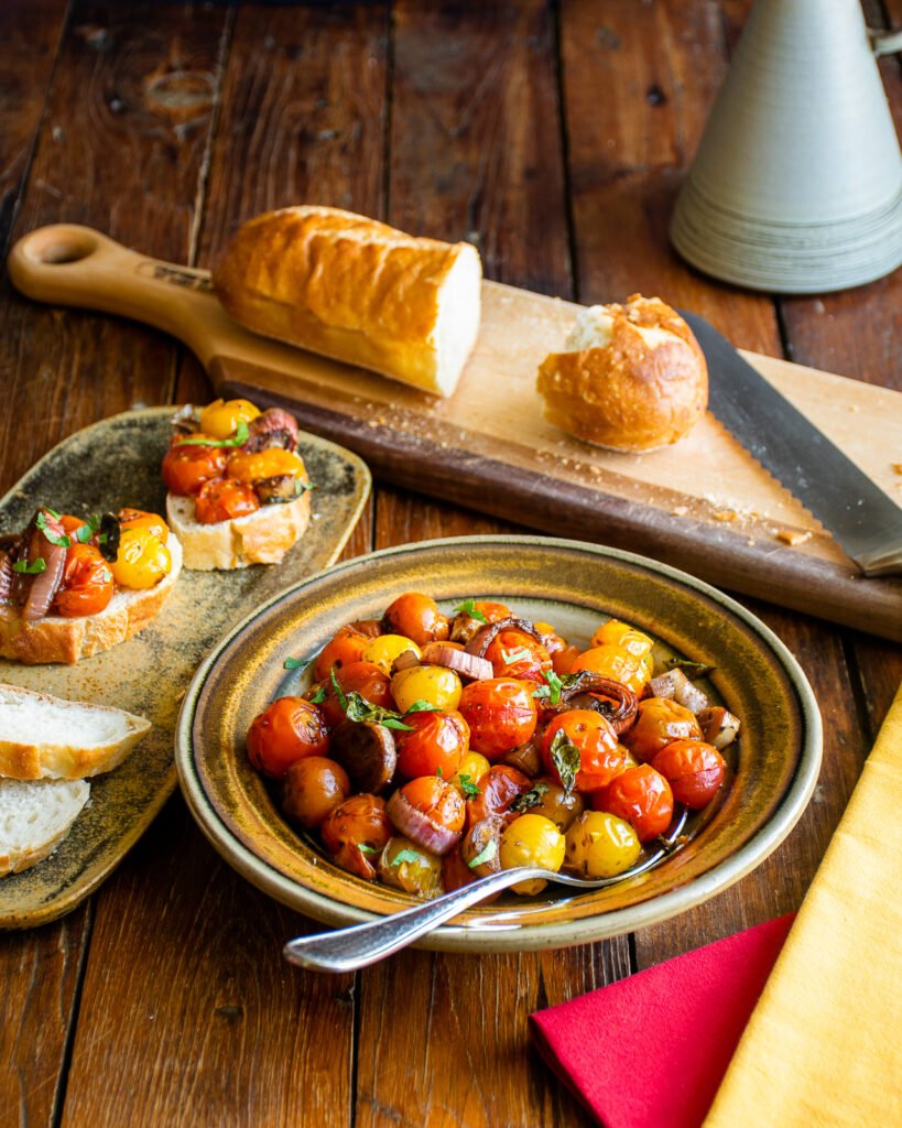 Roasted cherry tomatoes in a brown bowl with sliced baguette and crostini near by.