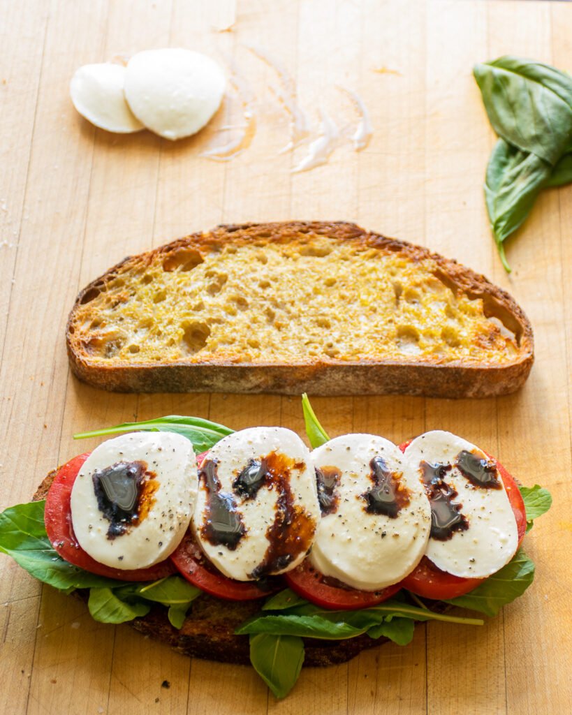 A sandwich being built on a wooden cutting board with fresh mozzarella and balsamic vinegar on top.