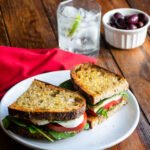 A Caprese Sandwich on a white plate with a red napkin, a glass of sparkling water and a ramekin of black olives on a wooden table.