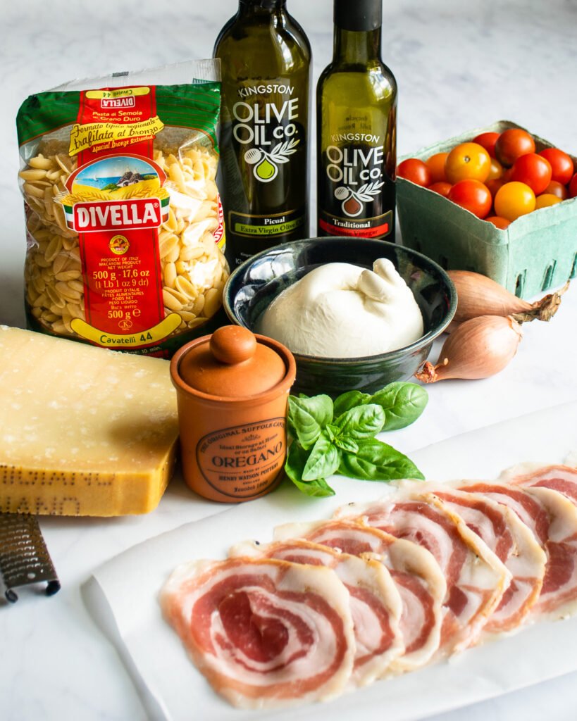 Ingredients on a counter - pancetta, parmesan cheese, oregano, pasta, burrata, cherry tomatoes, balsamic vinegar, olive oil, shallots and basil.