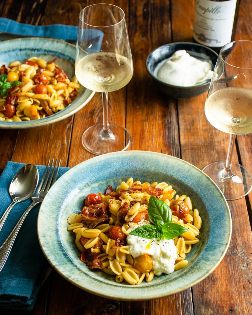 A blue bowl of cavatelli pasta with roasted cherry tomatoes, pancetta and burrata on a wooden table.