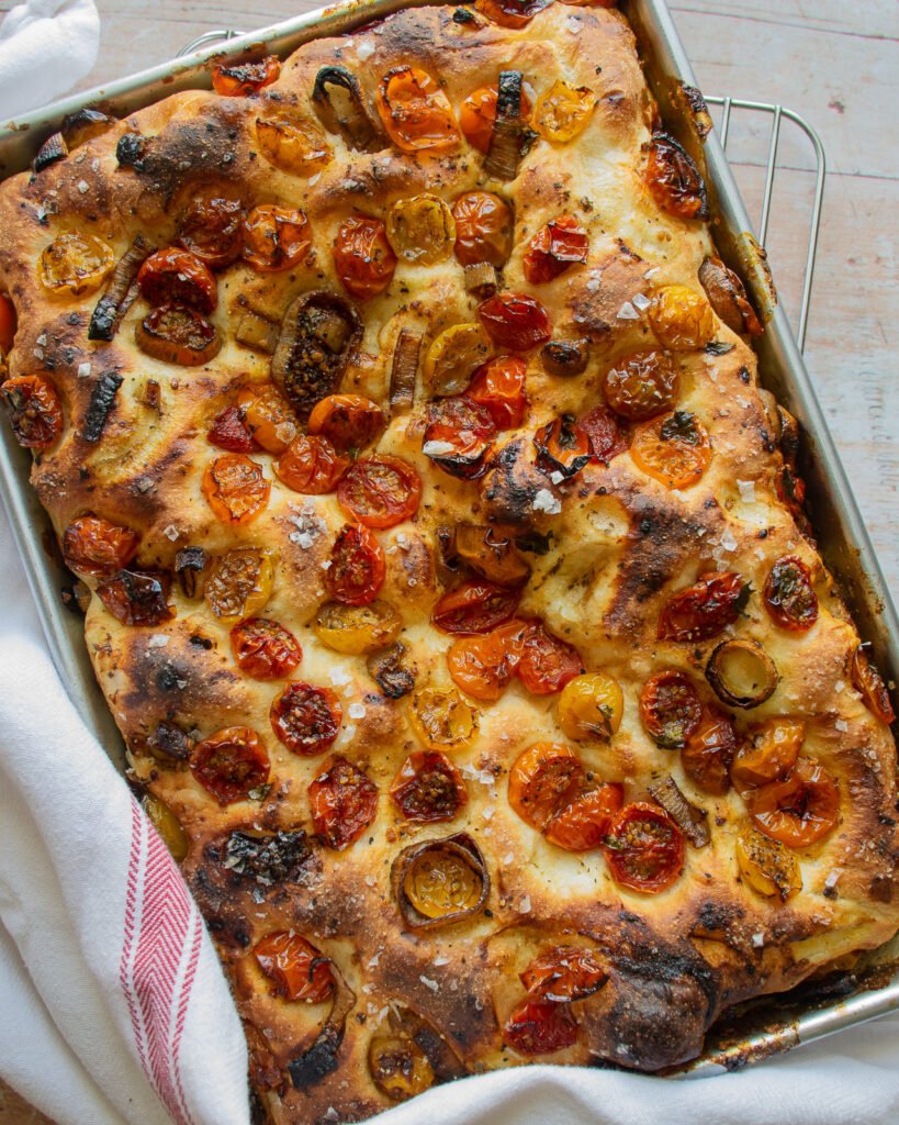 Roasted Cherry Tomato Focaccia bread in a baking pan on a cooling rack with a red and white striped towel.