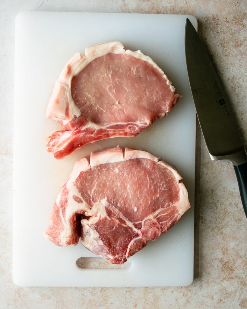 Two pork chops on a white cutting board with a knife next to it.