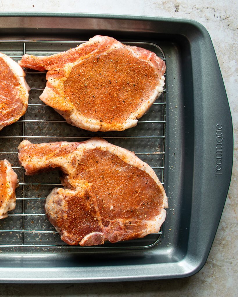 Seasoned pork chops on a rack on a baking tray.