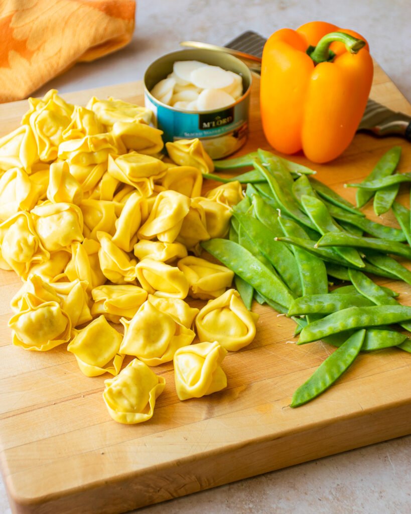 Ingredients on a cutting board - tortellini, snow peas, orange pepper and a can of water chestnuts.