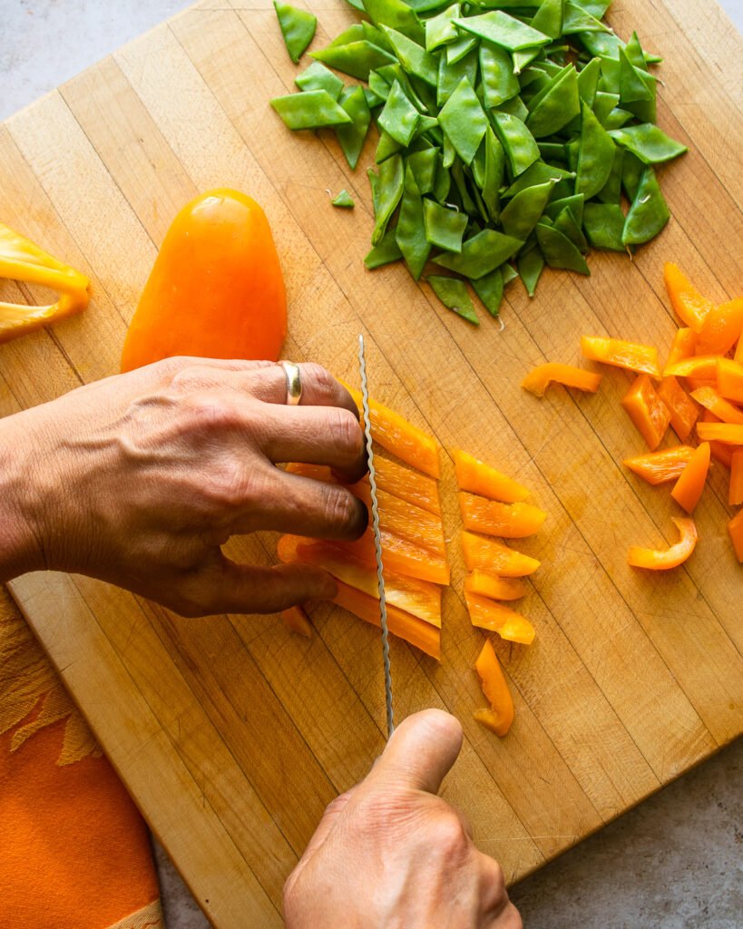 Hands cutting an orange pepper into pieces on a cutting board.