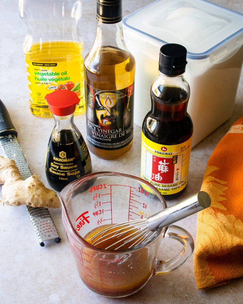 Ingredients on a countertop - soy sauce, sesame oil, vegetable oil, rice wine vinegar, fresh ginger and sugar - with a measuring cup of vinaigrette in front.
