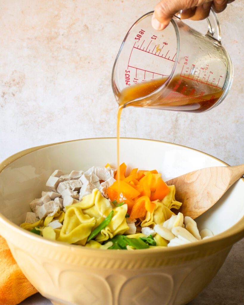 Pouring dressing on sesame chicken tortellini salad in a large bowl with a wooden spoon.