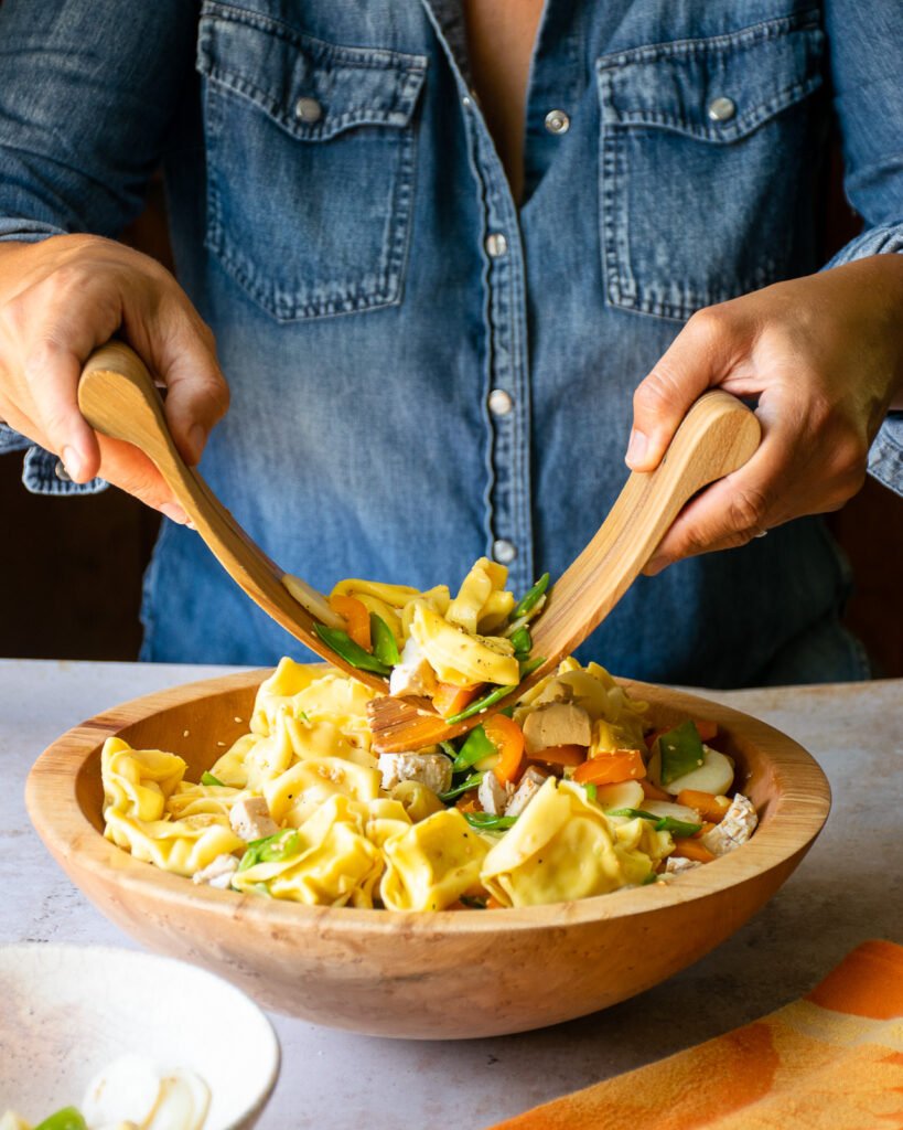 Two hands tossing sesame chicken tortellini salad with wooden servers.