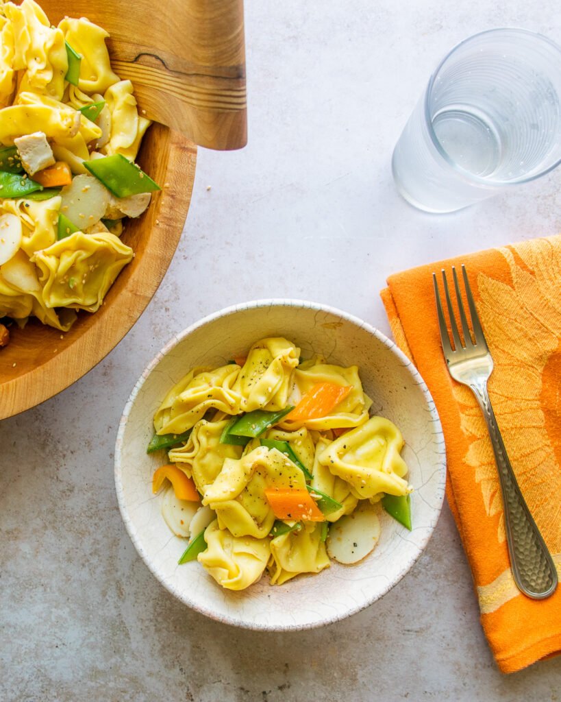 A serving of sesame chicken tortellini salad next to a wooden bowl with an orange napkin.
