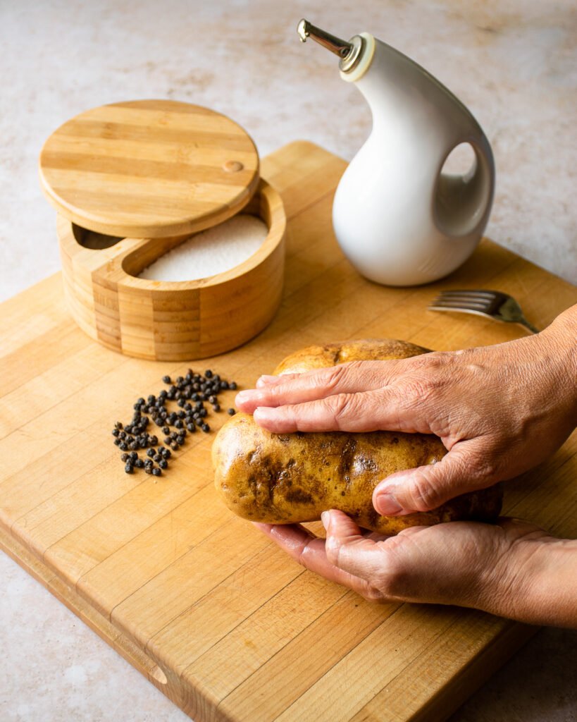 Two hands rubbing oil on a russet potato on a cutting board with salt, pepper and a cruet of oil near by.