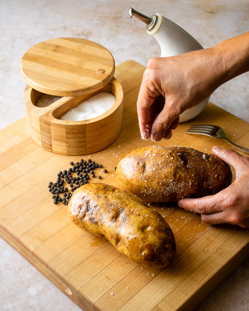 Hands seasoning russet potatoes with salt and pepper.