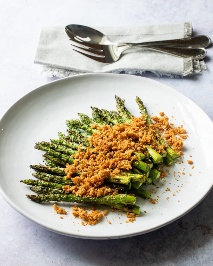Asparagus with crispy parmesan breadcrumbs on a white plate with serving utensils next to it.