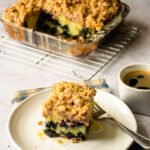 A piece of blueberry crumb coffee cake on a white plate with more behind and a blue checkered napkin.