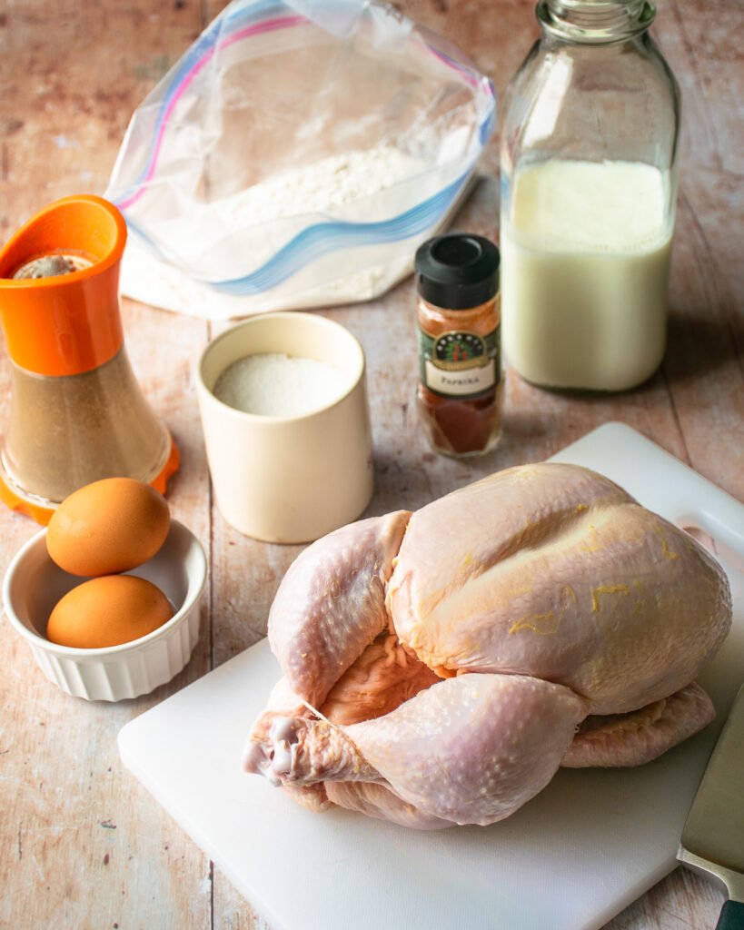 A whole raw chicken on a white cutting board with other ingredients around it.