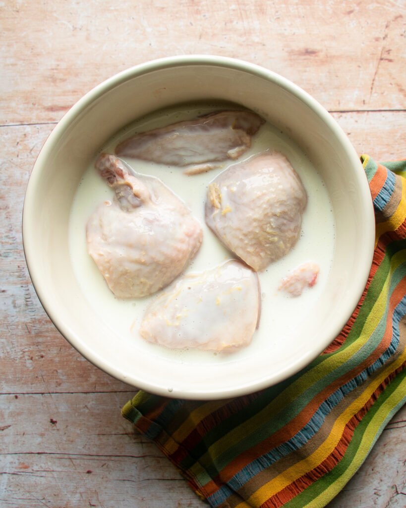 Chicken pieces soaking in buttermilk in a ceramic bowl on a wooden table with a striped tea towel near by.