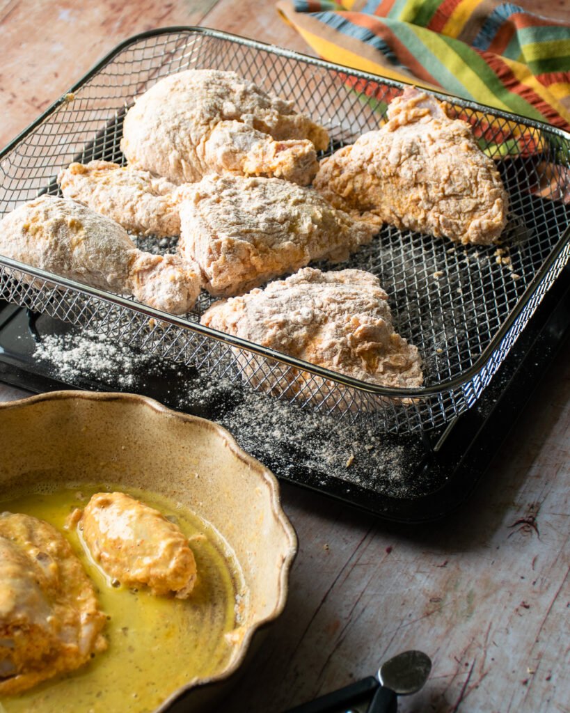 Dredged and coated chicken pieces in a mesh tray next to a shallow dish of beaten egg.