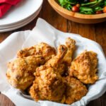 Buttermilk fried chicken in a parchment paper lined basket with a salad bowl in the background.